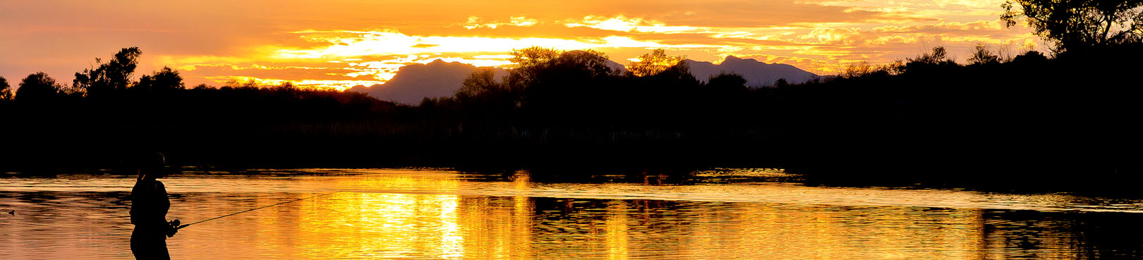 A person fishes on the shores of Patagonia Lake under an orange and gold sunset.