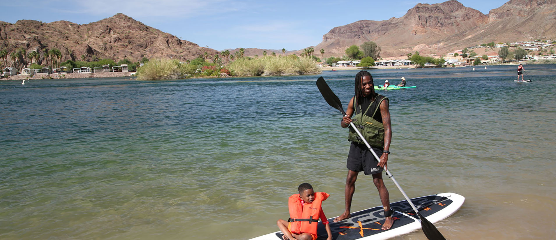 A man standing on a paddleboard with a child in a lifejacket sitting in front of him on the Colorado River