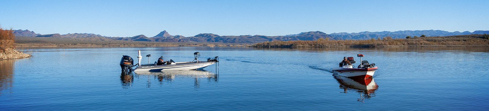 Two fishing boats on a still blue lake with mountains in the distance.