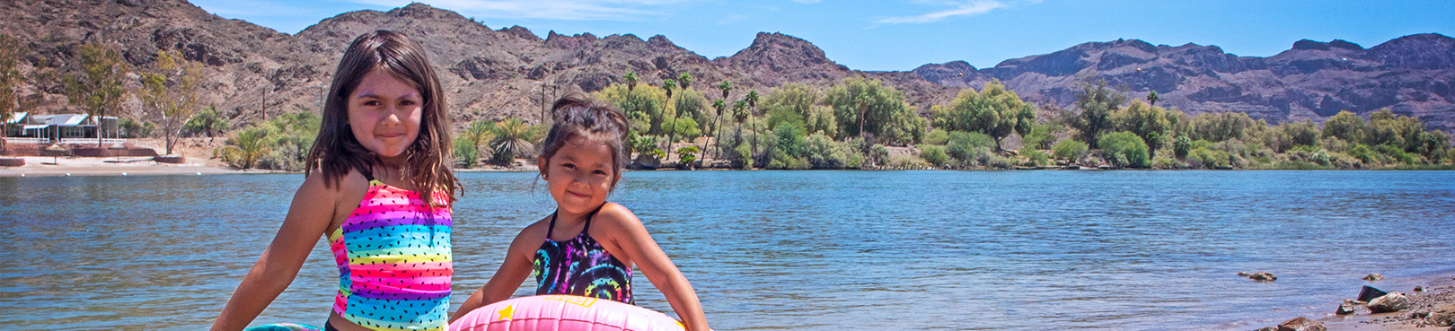 Two kids in swimsuits posing for a photo in front of the Colorado River with rugged desert mountains in the distance.