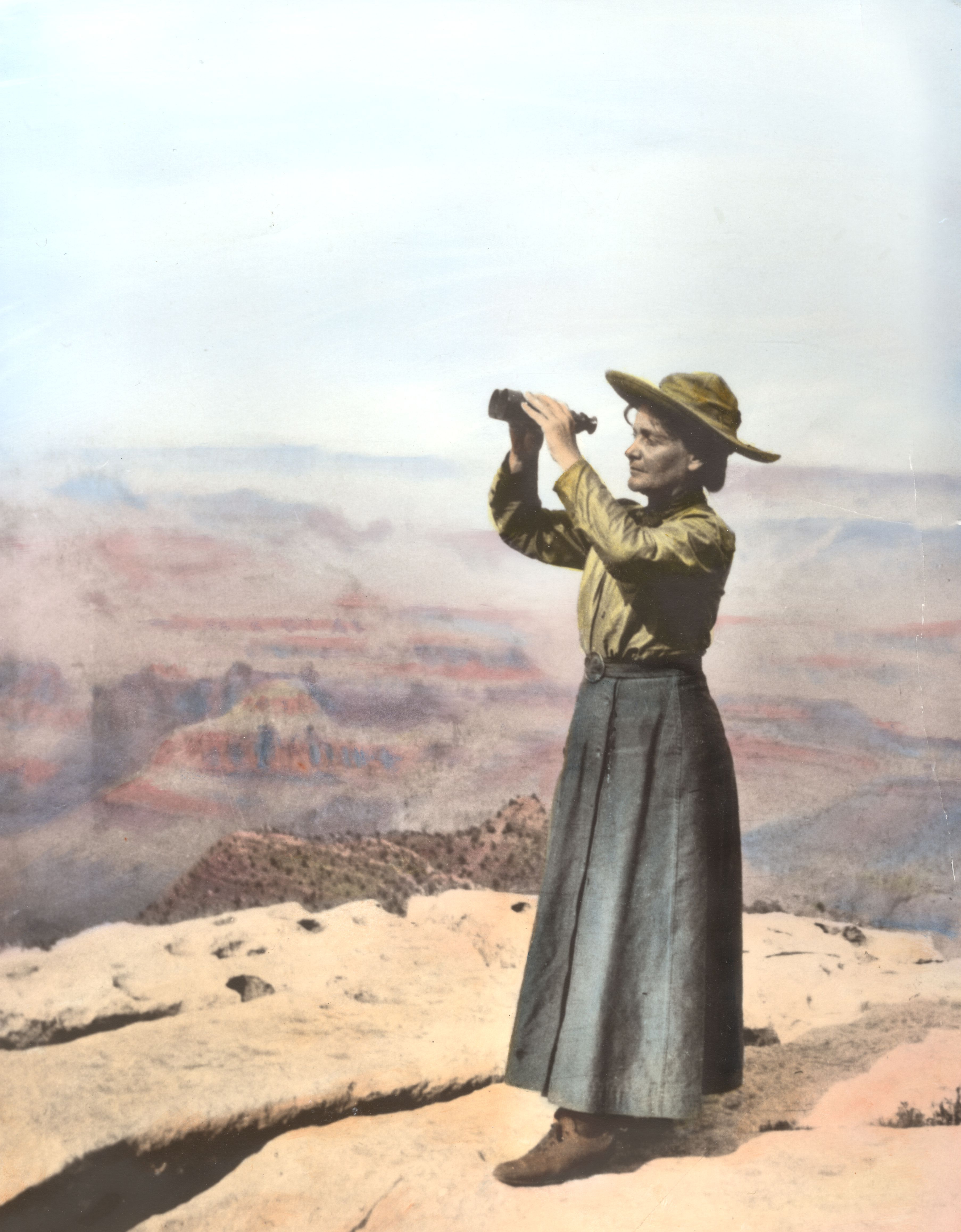 A vintage photo of Sharlot Hall in a dress holding up binoculars at the Grand Canyon.